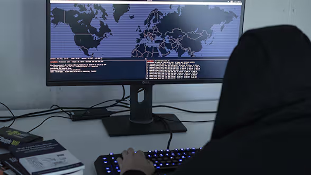 Photo symbolisant la cybercriminalité: assis devant un écran d’ordinateur montrant une carte du monde, un homme dont la tête est couverte d’une capuche modifie des codes de programmation.