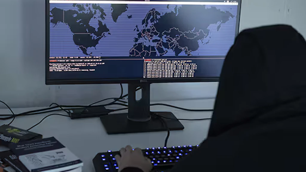 Photo symbolisant la cybercriminalité: assis devant un écran d’ordinateur montrant une carte du monde, un homme dont la tête est couverte d’une capuche modifie des codes de programmation.